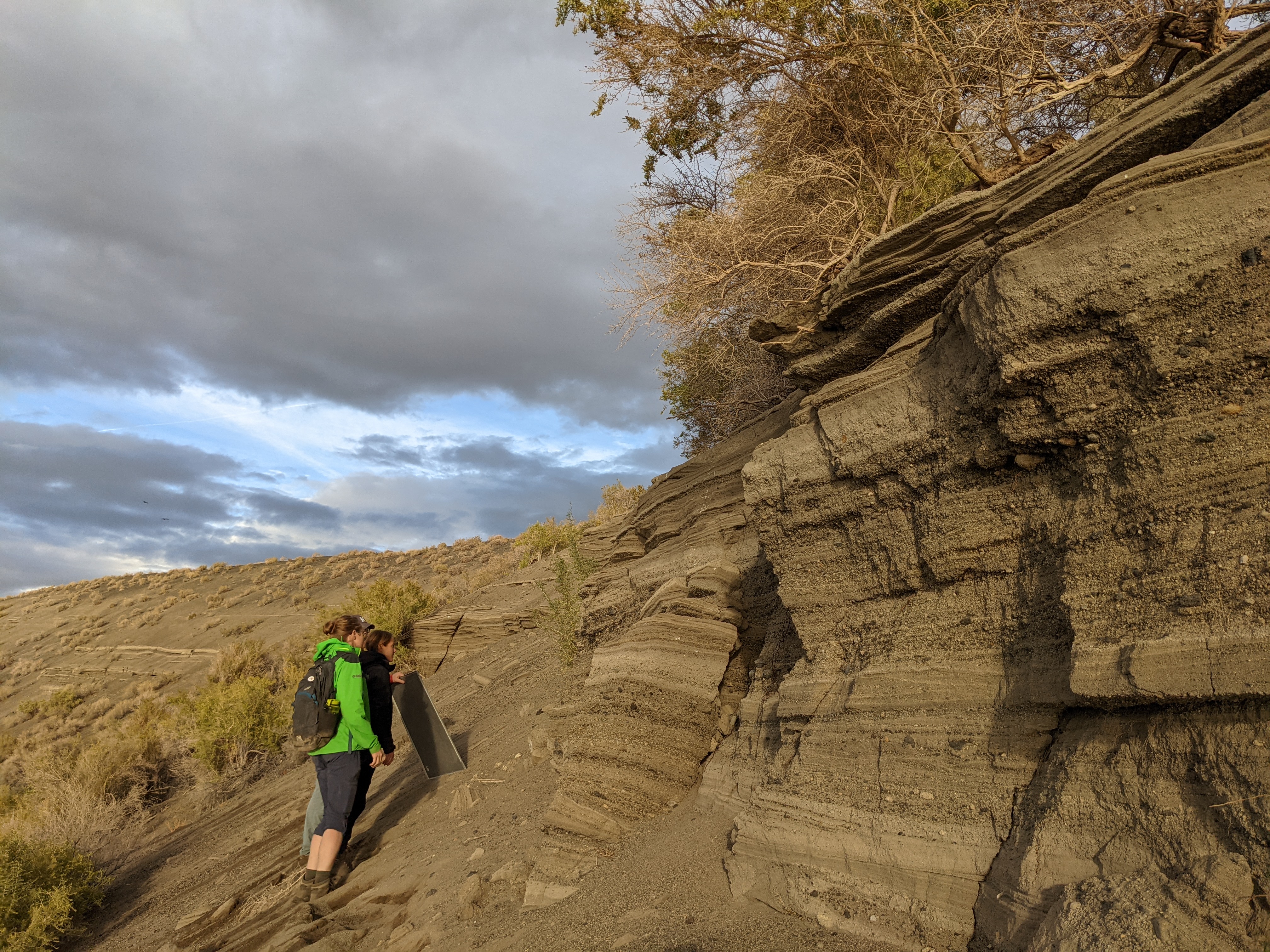 Three people examine tilted layers of volcanic ash and sediment near Soda Lake, Nevada. One holds a sample board. The outcrop is steep and textured, with dry shrubs and a cloudy sky in the background.